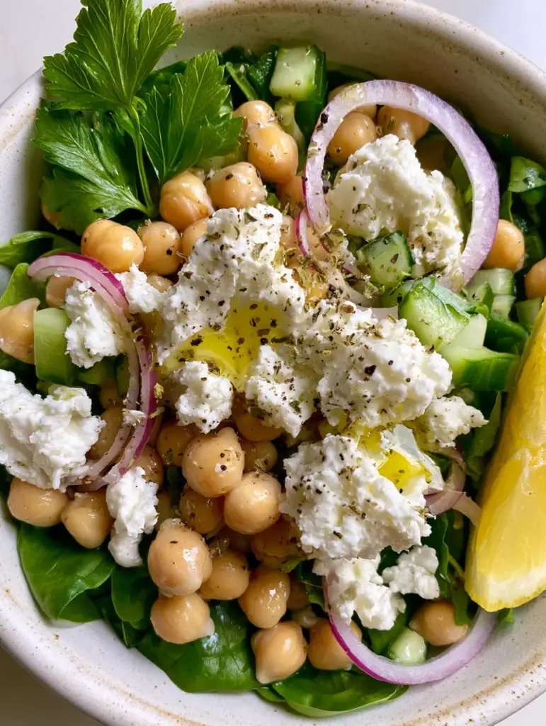 Top-down view of a rustic white bowl filled with spinach, chickpeas, cucumbers, and red onion drizzled with vinaigrette. (chickpea spinach feta salad)