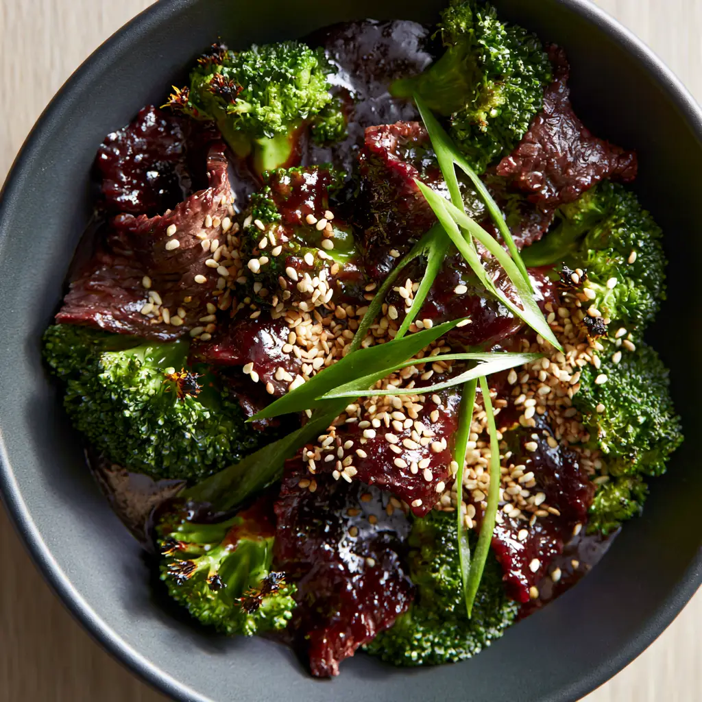 Top-down angled shot of beef and broccoli garnished with sesame seeds and scallions in a modern black bowl.