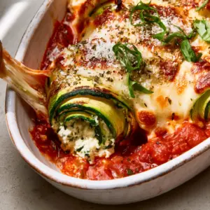 Close-up of Baked Zucchini Roll Ups showing charred zucchini ribbons, creamy ricotta filling, and toasted parmesan crust in a white ceramic dish.