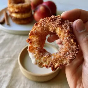 A hand holding a thick, golden-brown Air Fryer Cinnamon Apple Ring partially dipped in creamy white maple glaze.