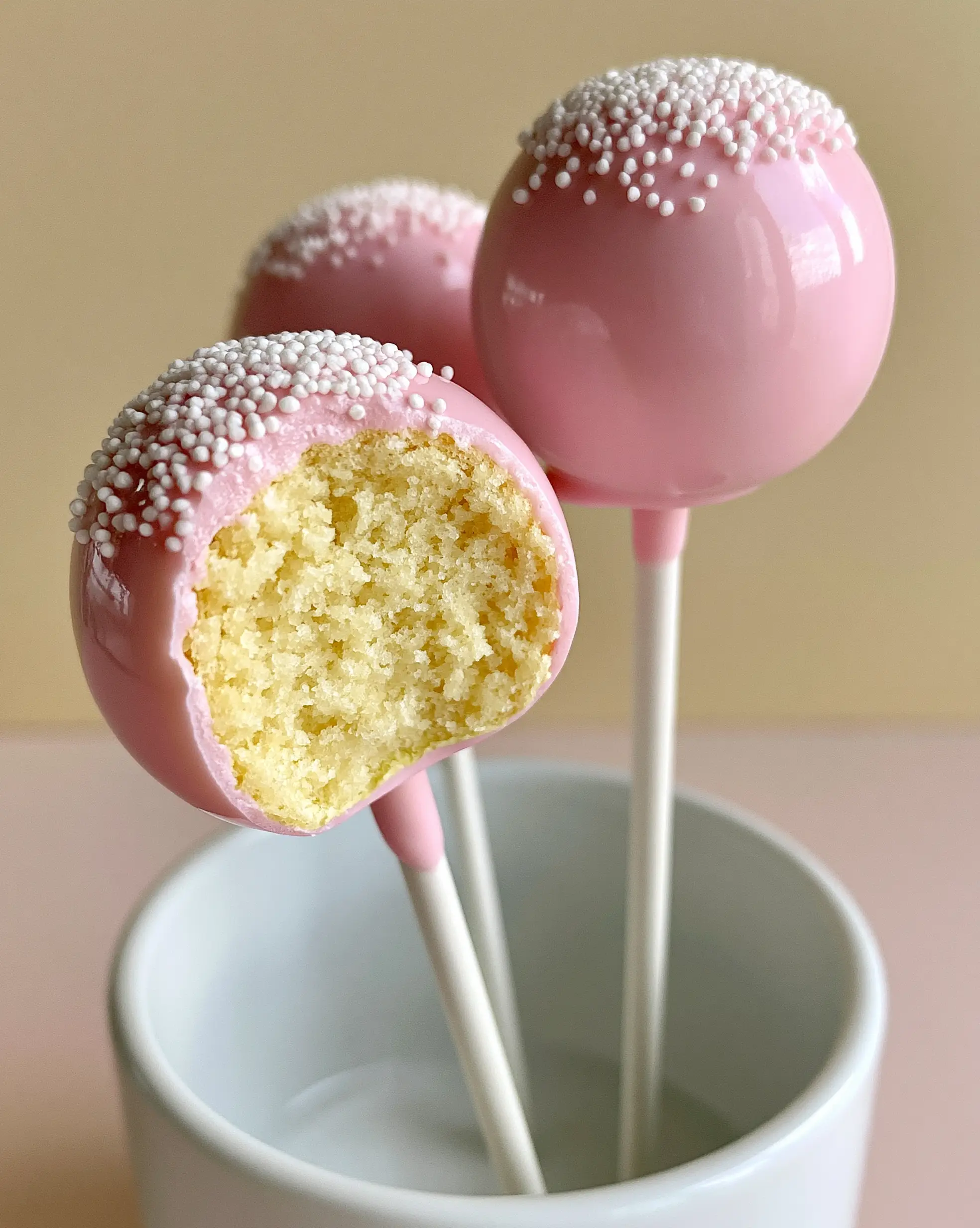 Three flawless pink cake pops standing in a ceramic holder, one bitten to show the moist yellow cake crumb. (cake pop recipes)